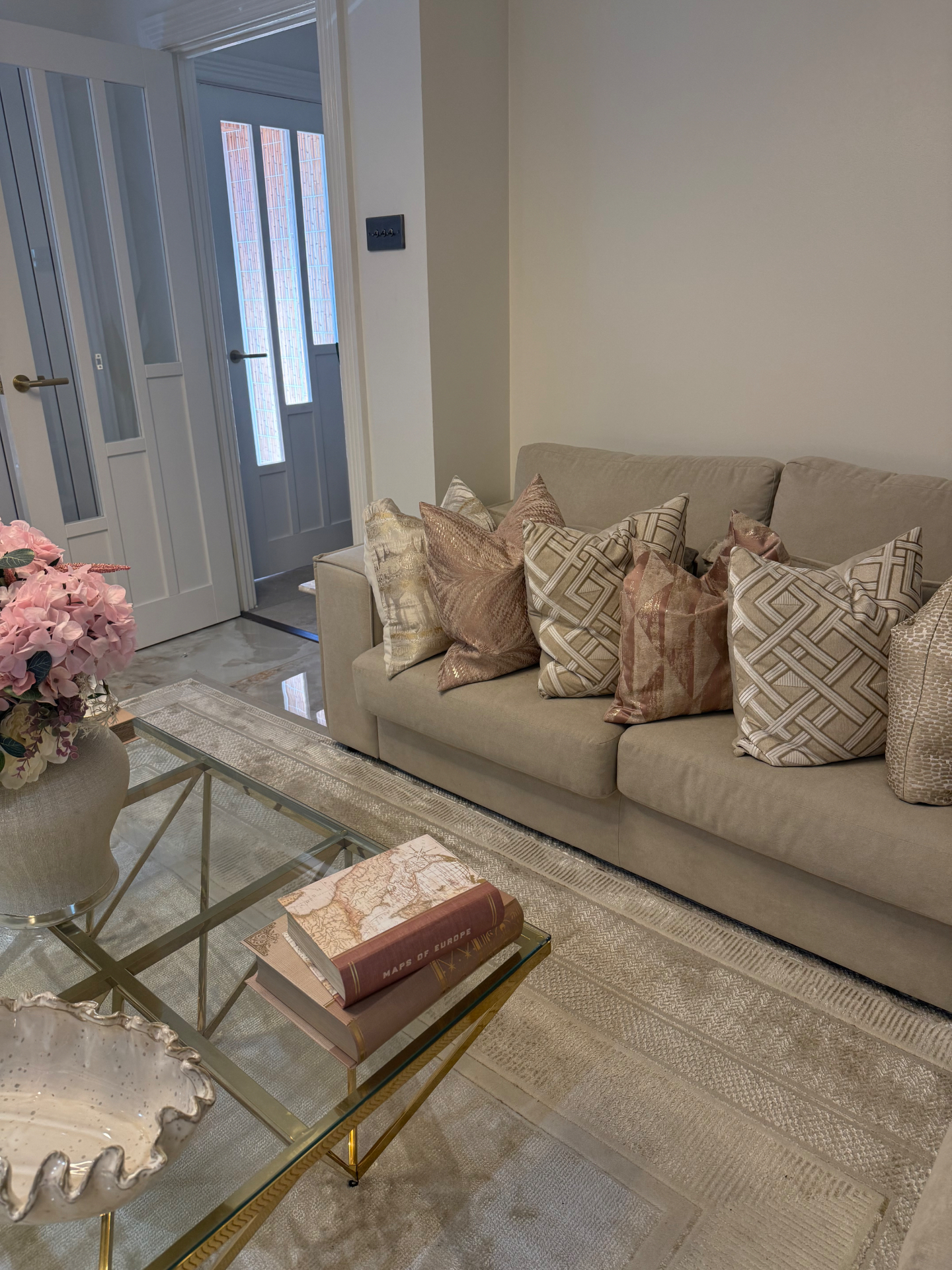 Neutral lounge with layered pink and geometric cushions, glass and brass coffee table styled with vintage map books